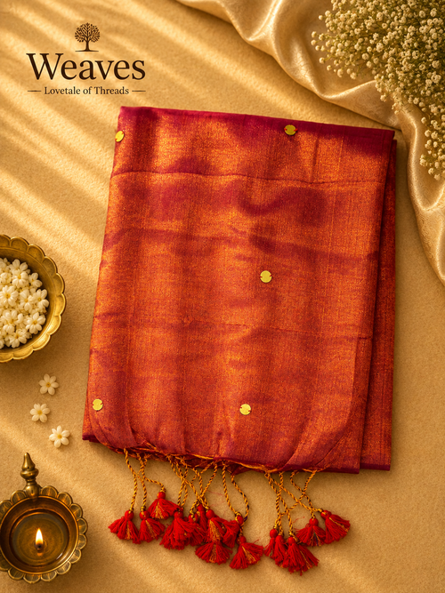 Burnt orange and crimson soft tissue saree with red tassels displayed on a warm golden backdrop